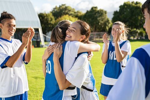 Team members in blue and white uniforms hug on a green field, others clap.