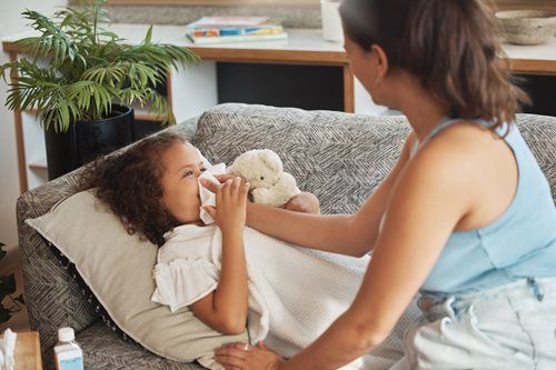 Child on couch blowing nose, being cared for by an adult. Interior, daytime.