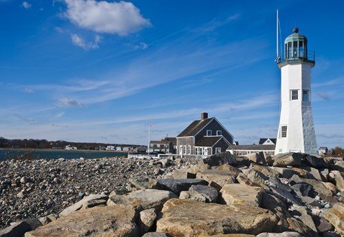 White lighthouse on a rocky coast, next to a gray building, under a blue sky.