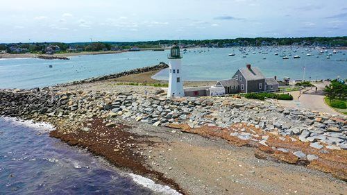 Lighthouse on a rocky breakwater, with a building, and boats in the background. Blue water and sky.