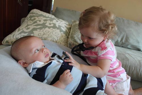 Baby lying on pillow; toddler listens with a stethoscope.