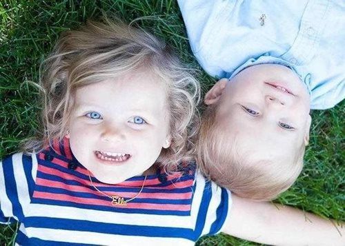 Two young children smiling, lying in grass; girl with blue eyes and striped shirt, boy with light hair.