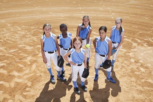 Softball team in light blue jerseys and white pants stand on a brown dirt field, posing with gloves and a softball.