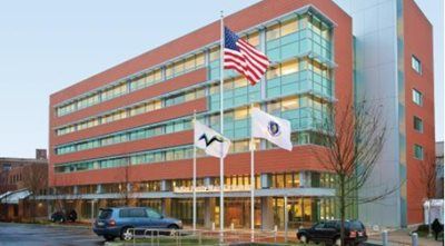 Modern brick and glass building with American, Massachusetts, and logo flags; cars parked out front.