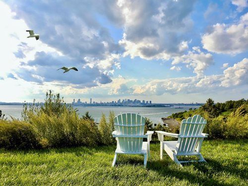 Two white Adirondack chairs on a grassy hill overlooking water and a city skyline under a cloudy blue sky.