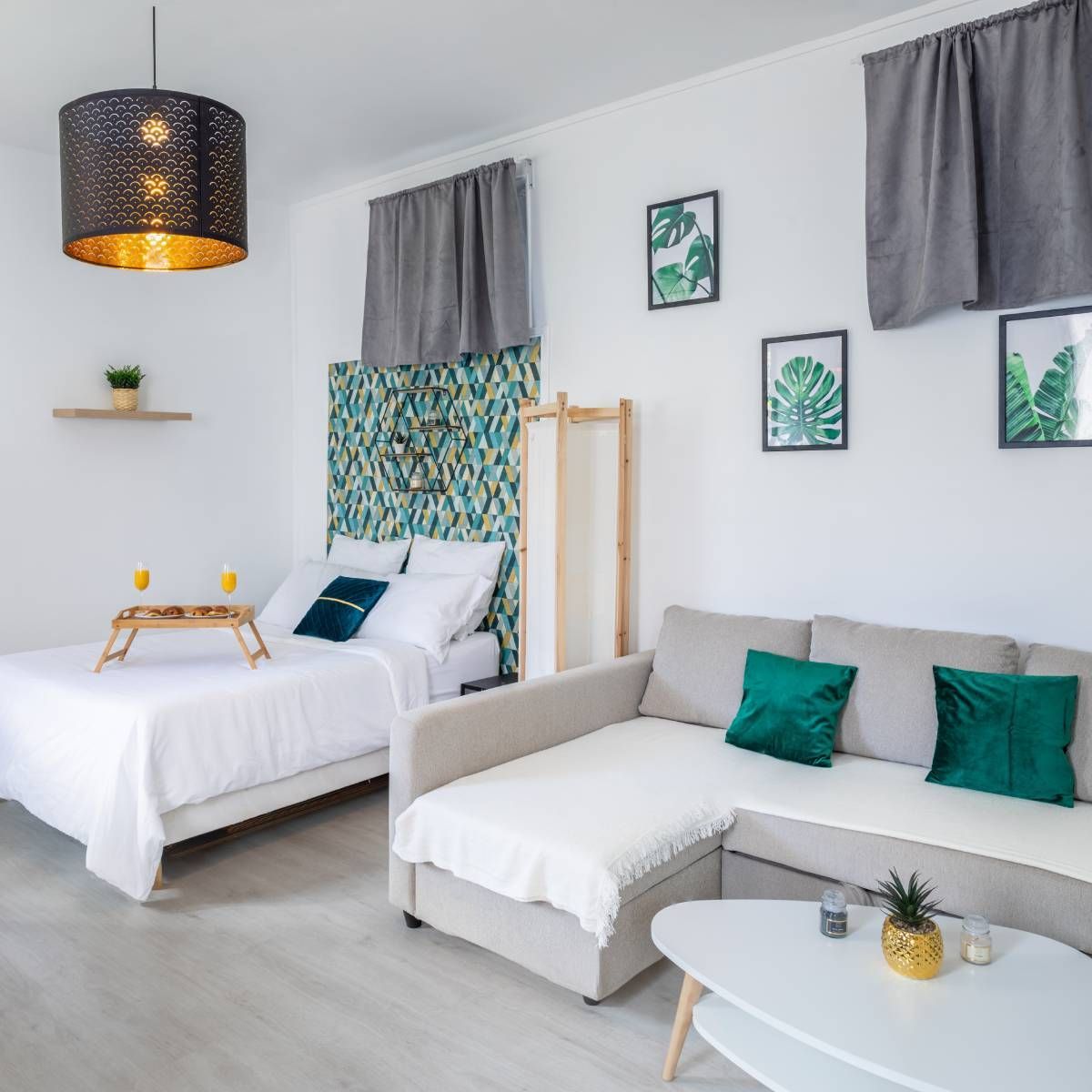 A bright studio apartment with a double bed, a grey sectional sofa, white decor, tropical leaf art, and a gold lamp.