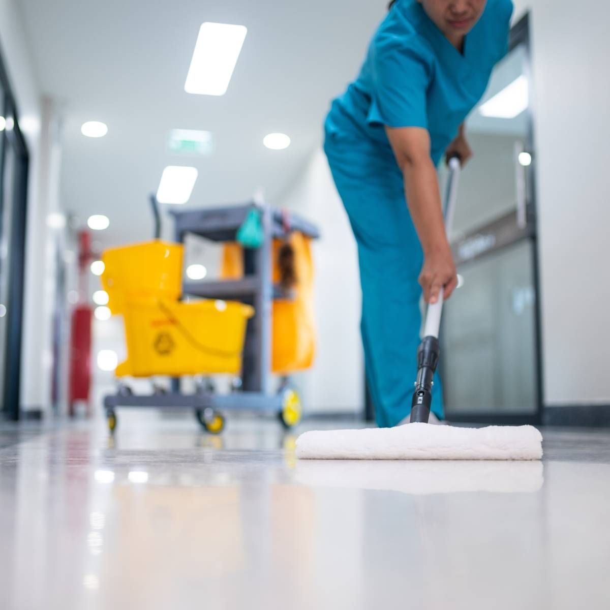 A person in teal scrubs mops a shiny floor in a brightly lit hallway, with a yellow mop bucket and cart in the background.