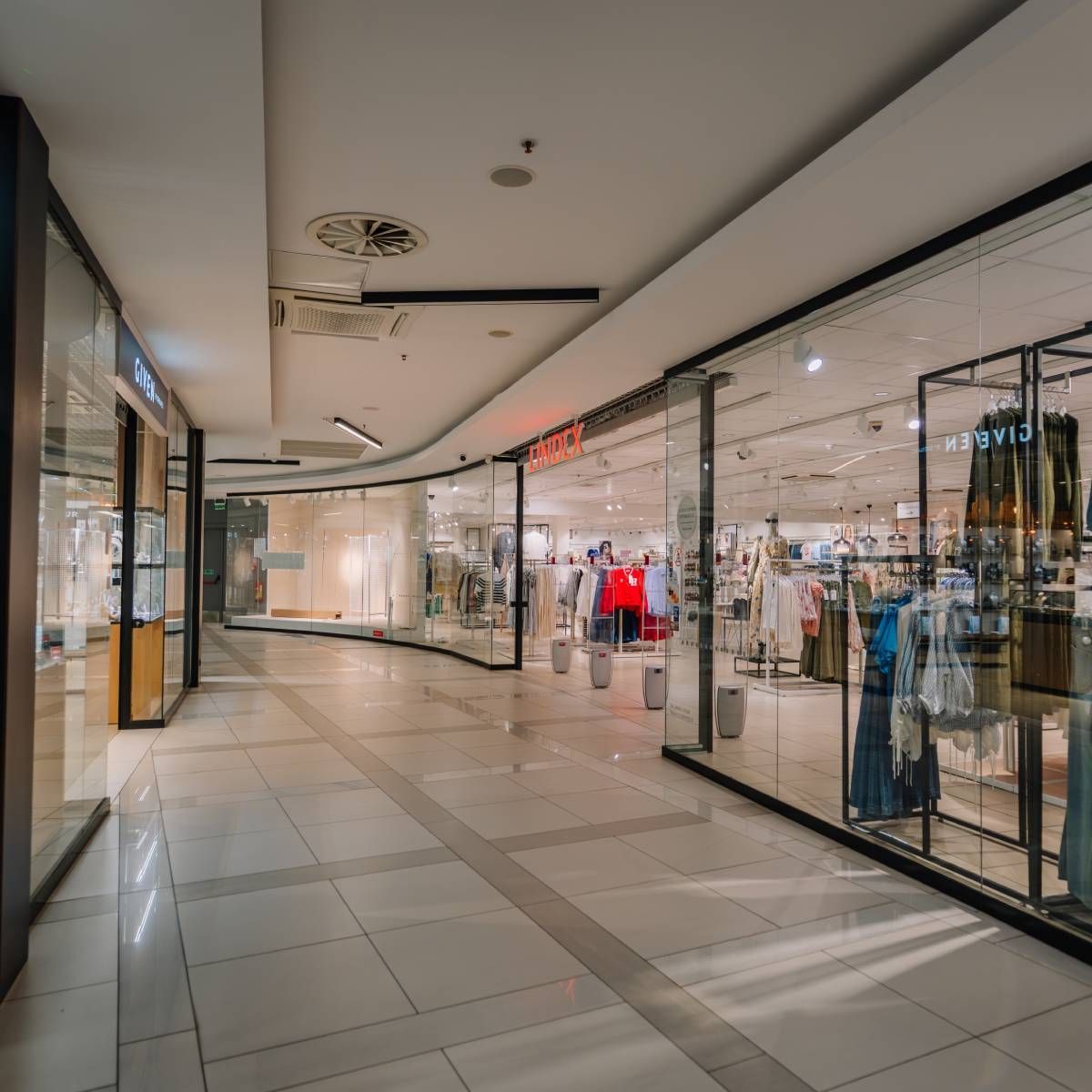 An empty corridor in a modern, brightly lit shopping mall with glass-fronted retail stores on both sides.
