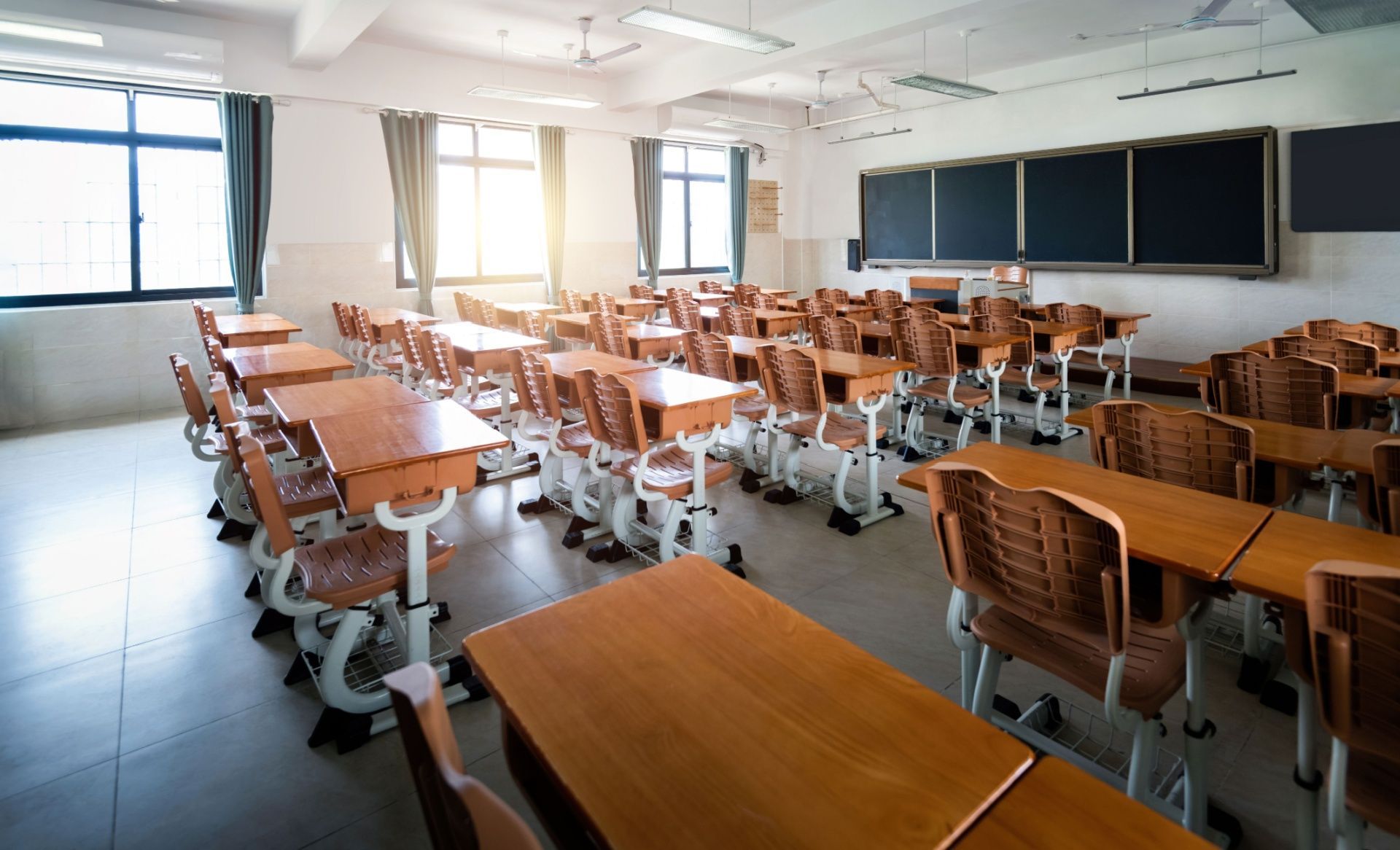 Empty classroom with wooden desks, chairs, and chalkboards near large windows and bright sunlight