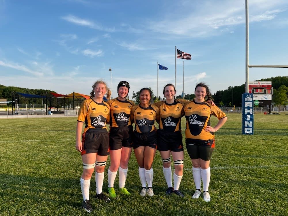 Five rugby players in yellow and black jerseys stand side-by-side smiling on a grassy field under a bright blue sky.