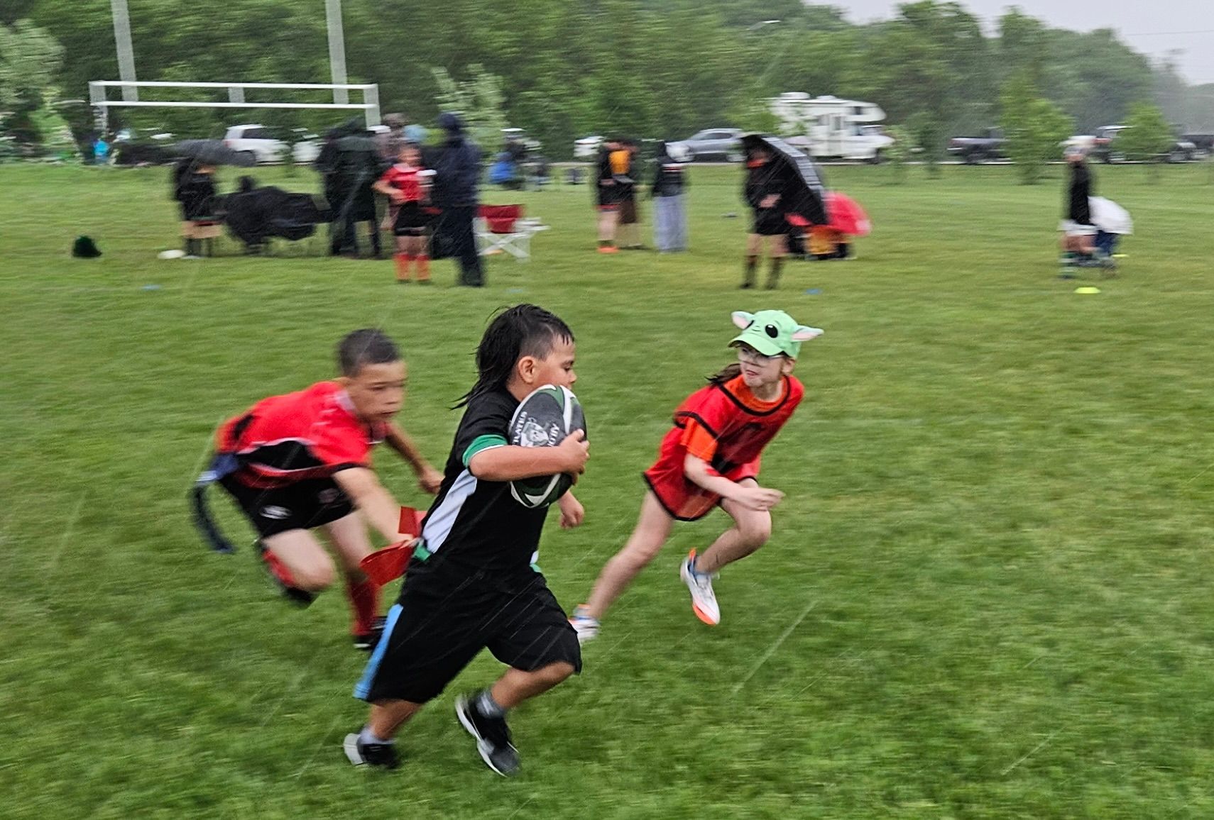 Three children play flag rugby on a grassy field in the rain, with spectators and a goal post in the background.