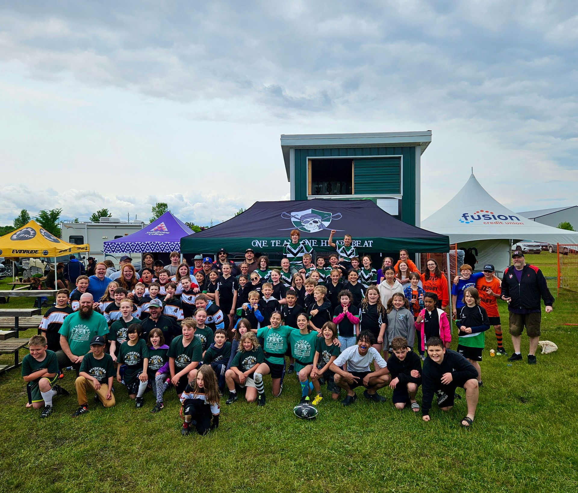 A large group of youth rugby players pose for a photo in front of tents at an outdoor field after a youth rugby jamboree.