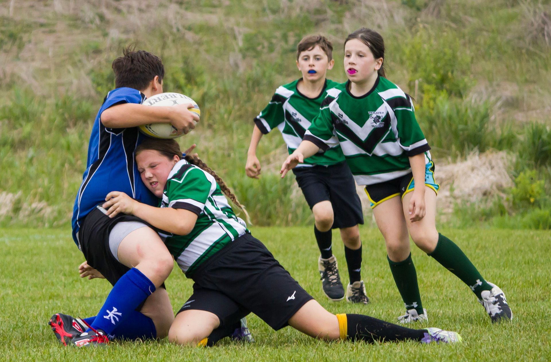 Players in blue and green uniforms compete during a rugby match on a grass field, with one tackling a ball carrier.