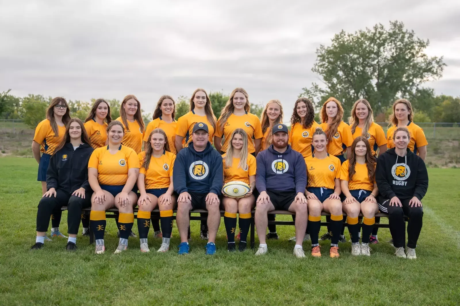 A group of rugby players in yellow jerseys and navy shorts pose for a team photo on a grassy field with two coaches.