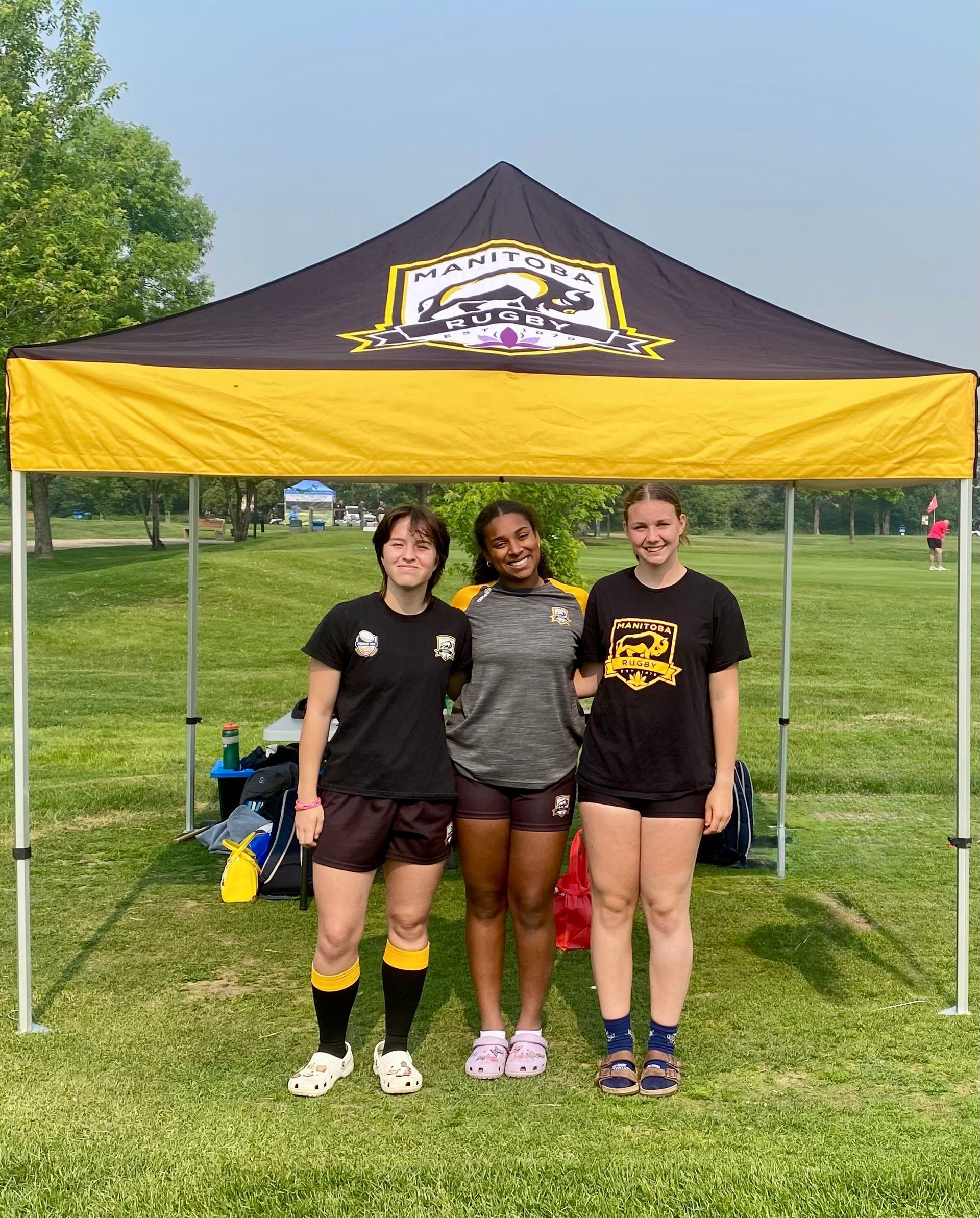 Three people stand smiling under a black and gold event canopy in a grassy park on a sunny day.