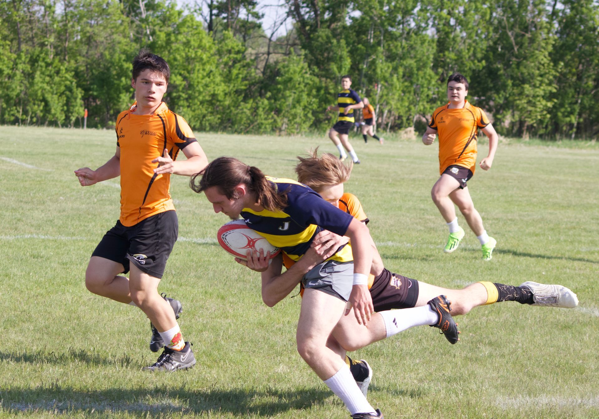 Rugby players in orange and blue-and-yellow uniforms compete on a sunny grass field.