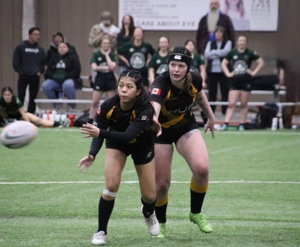 Two players in black and gold uniforms pass a rugby ball during an indoor match, with spectators in the background.