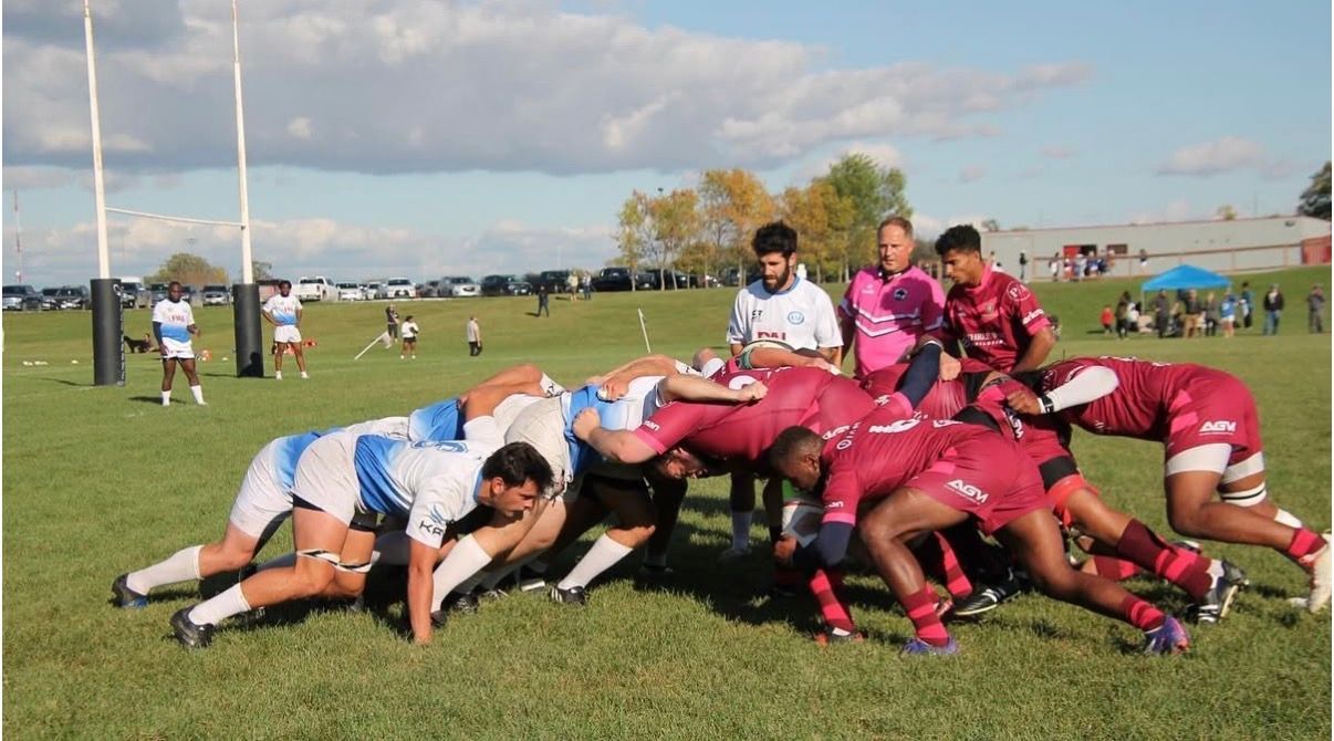 A rugby team in blue and white jerseys locks into a scrum against a team in maroon jerseys on a sunny outdoor field.