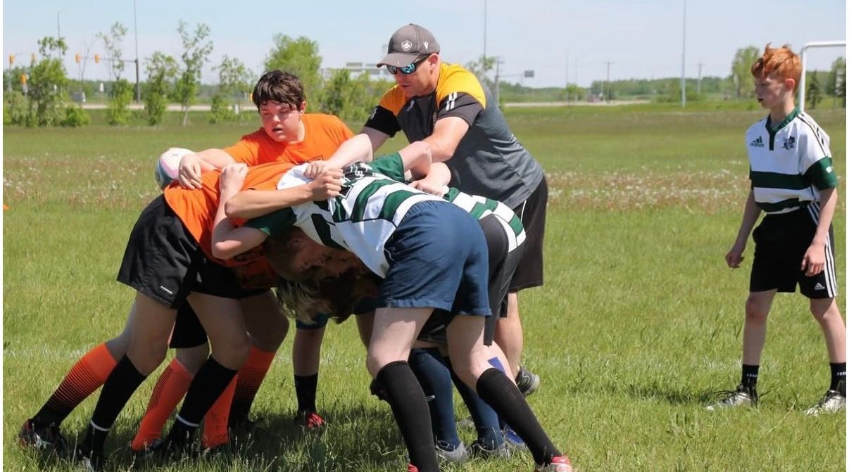 Players in orange and green jerseys engage in a rugby scrum on a grass field while a coach watches nearby.