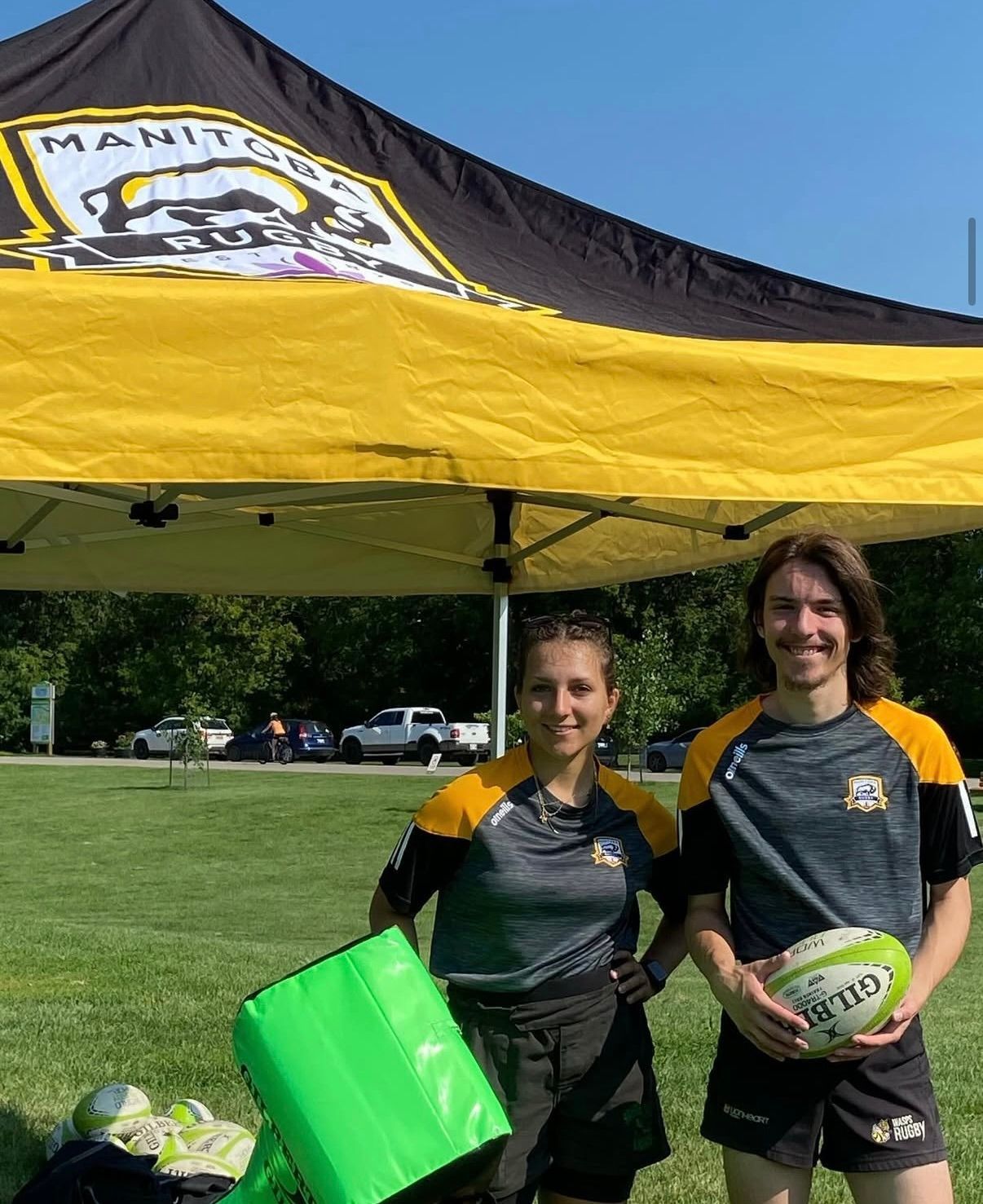 Two people in athletic gear hold a rugby ball under a Manitoba Rugby canopy on a sunny, grassy field.