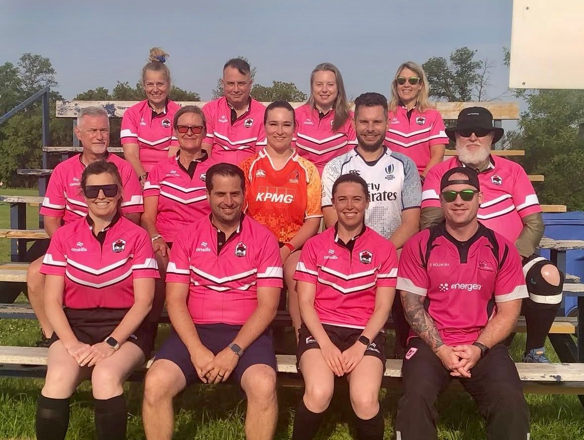 A group of match officials posing on outdoor bleachers, wearing matching pink sports jerseys, with several members wearing sunglasses.