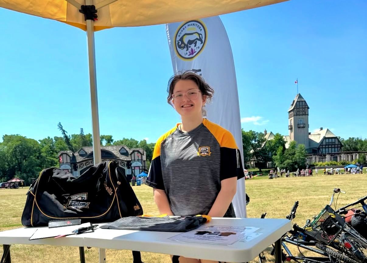 A person stands at an outdoor registration table in a grassy field, with a historical building and blue sky in the background.