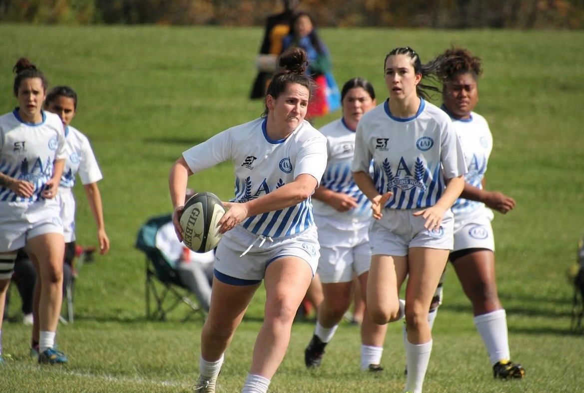 Players in white and blue striped uniforms run across a grass field, with one athlete in the foreground holding a rugby ball.