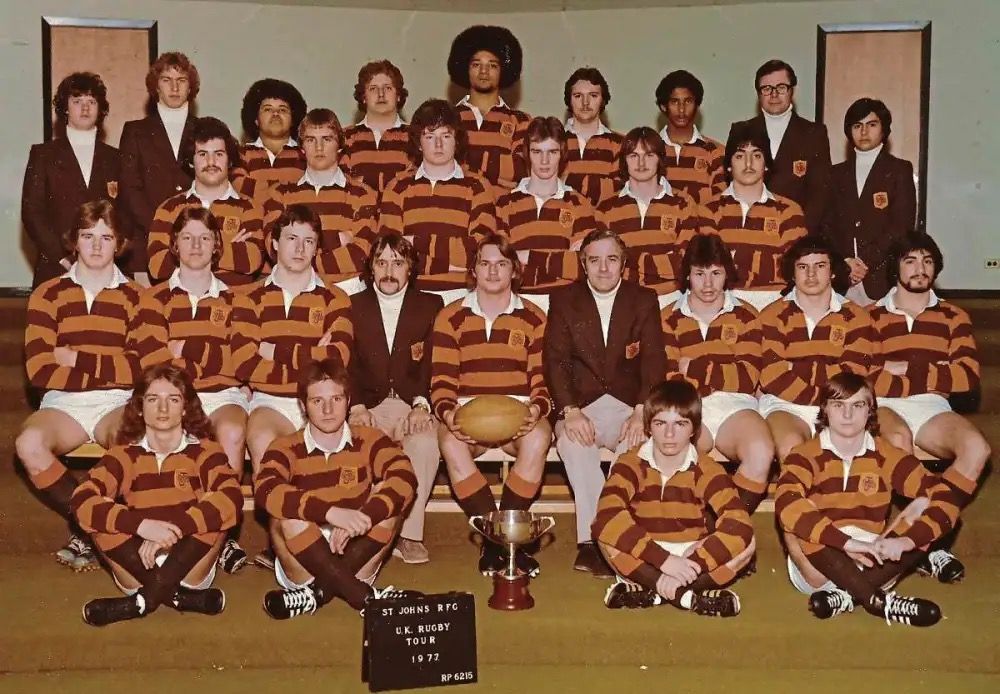 A rugby team posing in brown and gold striped jerseys with a trophy and sign in front, indoors.