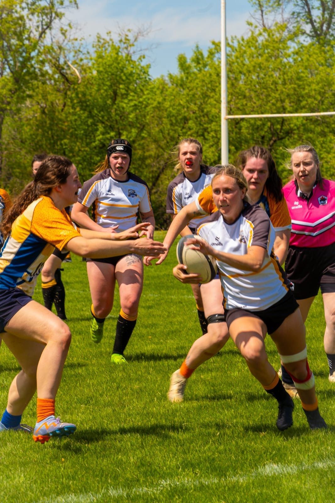 Rugby players in white and yellow jerseys on a sunny field, with one player carrying the ball and another reaching out.