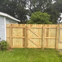 Wooden fence with gate, light brown, in front of a house, set in grass.