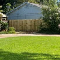 A green lawn leads to a wooden fence and a light blue house. Trees and bushes in the background.