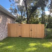Wooden fence in a grassy yard next to a brick building, under a blue sky with trees.