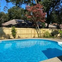 Pool surrounded by wooden fence, trees, and house under a blue sky.