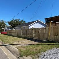 Wooden fence surrounds a house with blue siding on a sunny day. Green grass and blue sky.