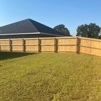 Wooden fence surrounding a grassy yard with a house and clear blue sky in the background.