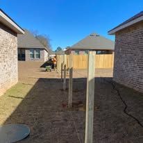 Wooden fence posts installed on a grassy area between two brick buildings under a blue sky.