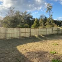 Wooden fence in a backyard with dry grass, trees, and a cloudy sky.