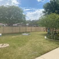 Backyard with wooden fence, green grass, and trees under a blue sky.