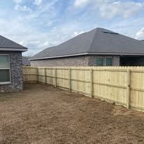 Wooden fence surrounding a backyard with a house in the background. Brown yard and cloudy sky.