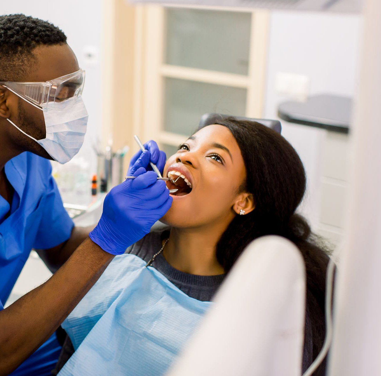 Black dentist examining a Black woman's teeth in a dental chair. Both wear masks and gloves.