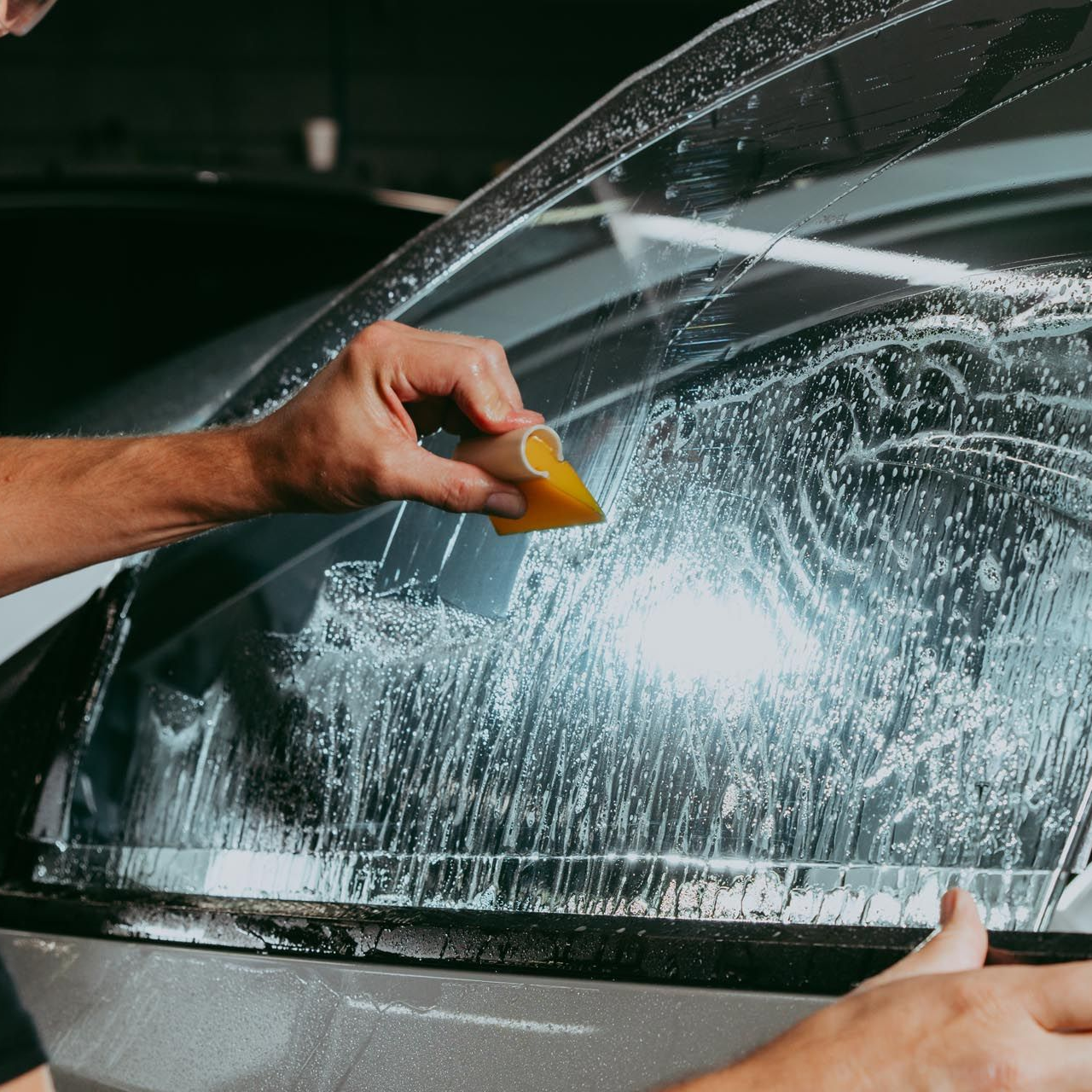 Person applying film to a car windshield with a yellow squeegee.