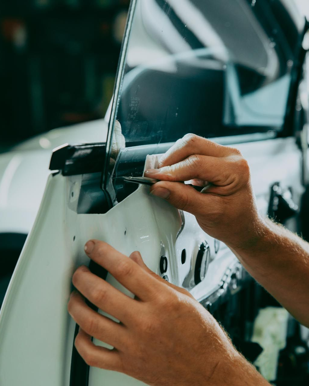 Hands working on a car door, using a tool on a window. Workshop setting.