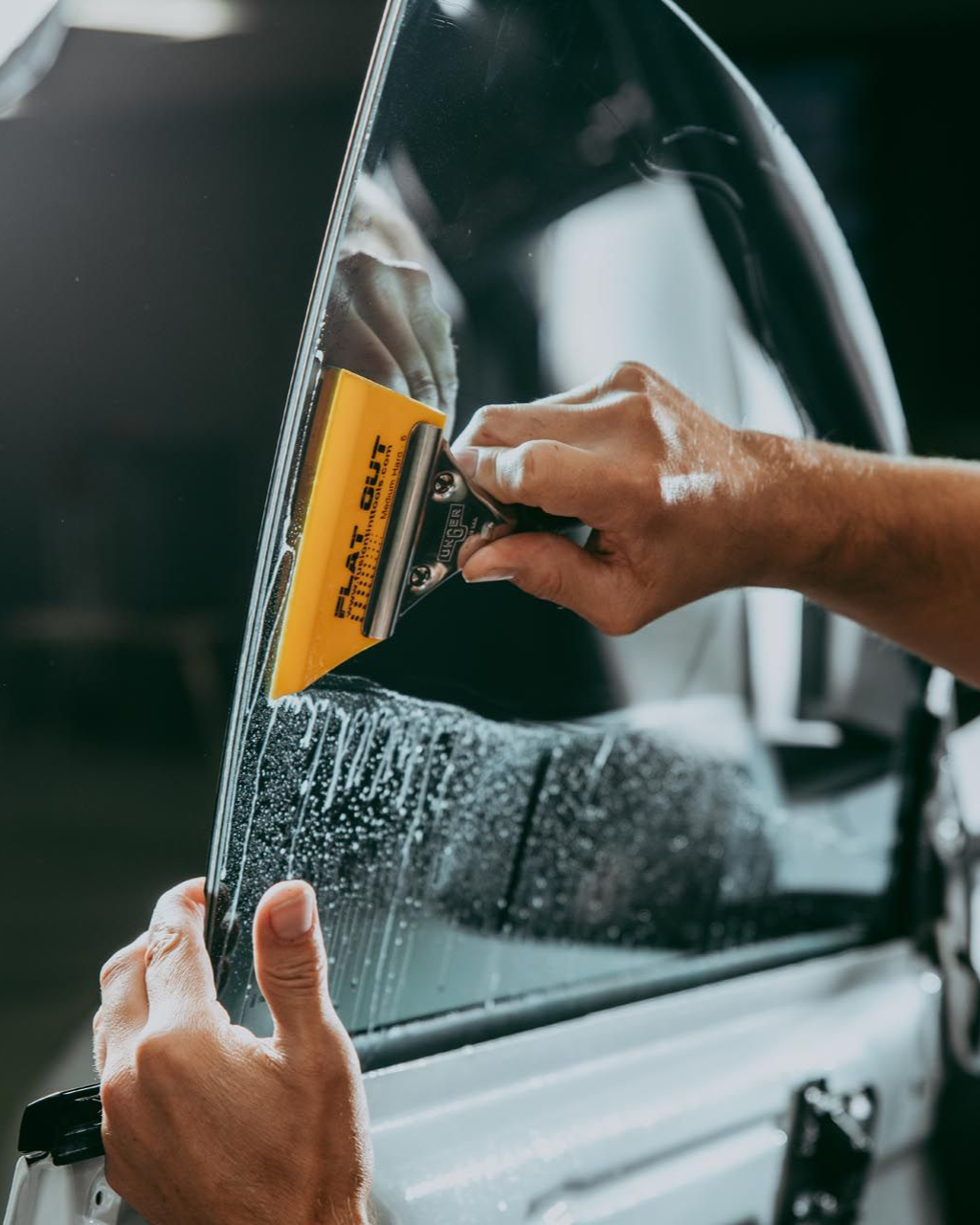 Person applying window tint to a car window with a yellow squeegee.