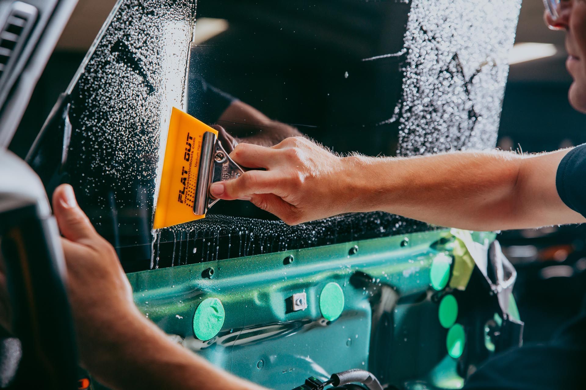 Person applies window tint to a car window with a yellow squeegee.