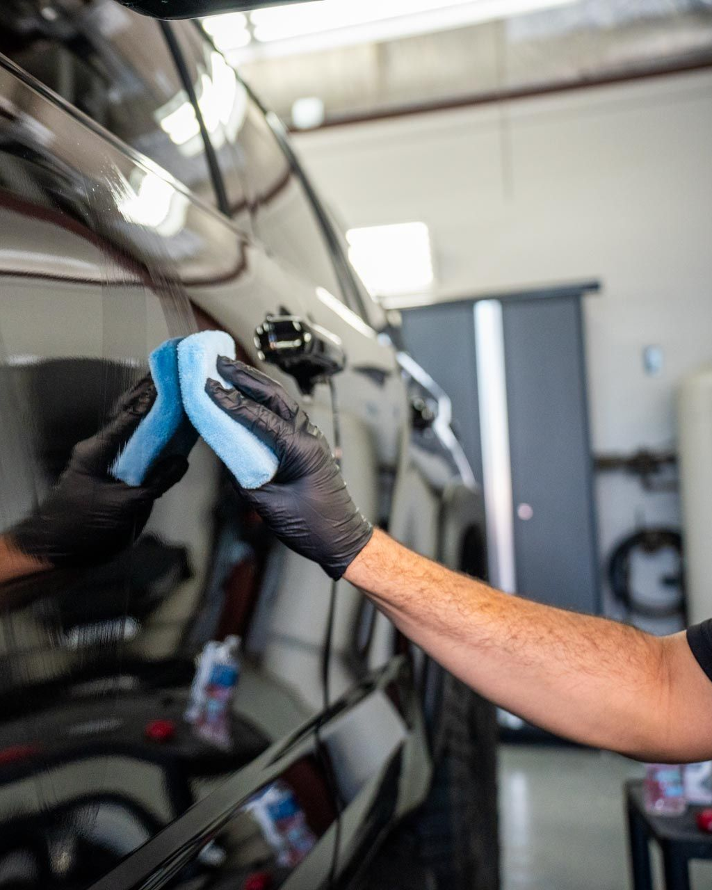 A person wearing black gloves polishes a black car with a blue sponge.