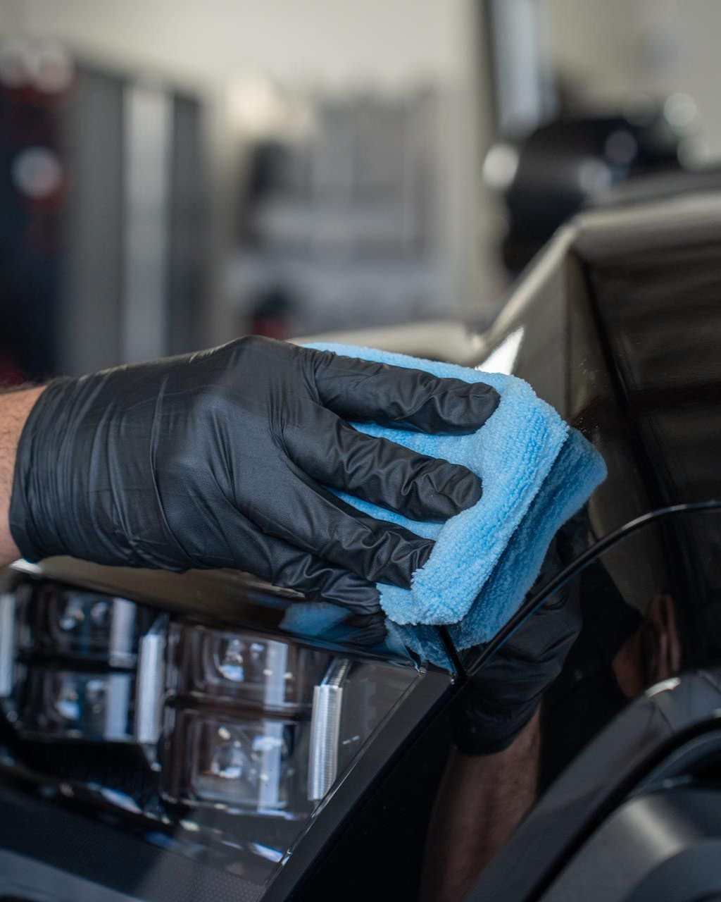 Black-gloved hands wiping a car's black surface with a blue microfiber cloth, in a shop setting.