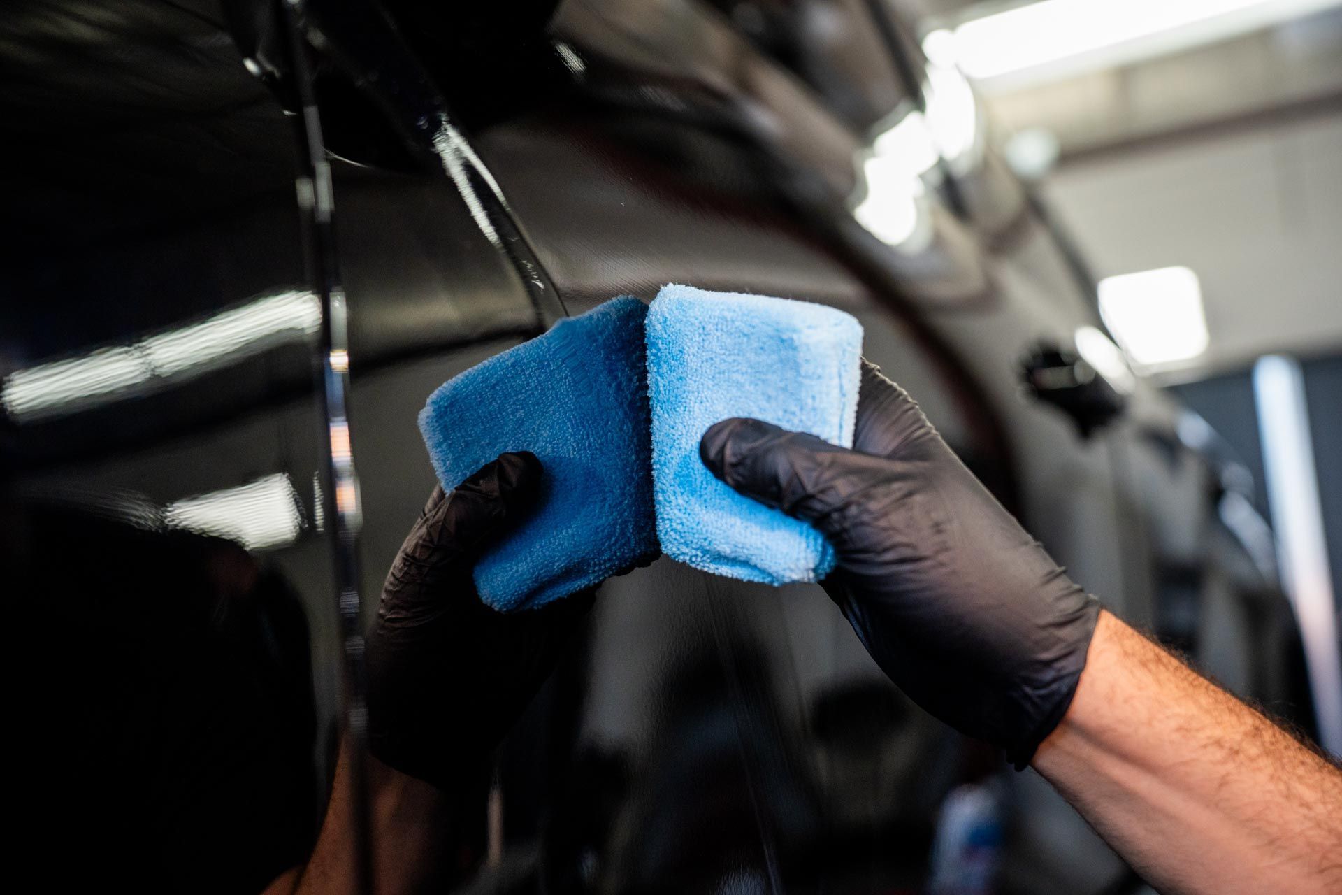 Gloved hands polishing a black car with two blue foam applicators in a garage.