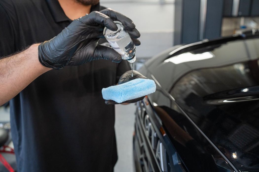 Person in black gloves applying liquid from a bottle onto a blue applicator pad on a black car.