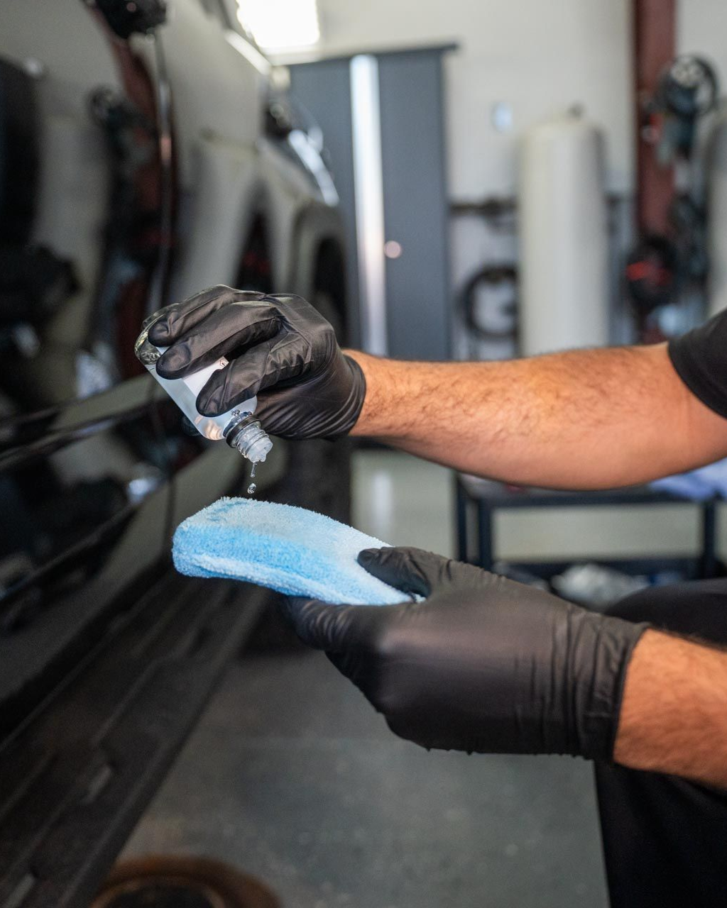Person wearing black gloves applying liquid from a bottle onto a blue applicator pad near a black car.