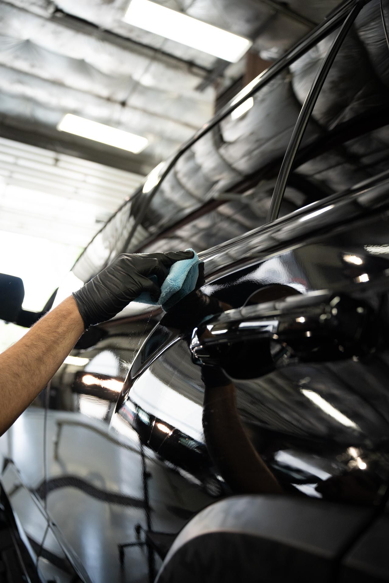 Gloved hand applying a blue detailing pad to a black car's surface in a brightly lit garage.
